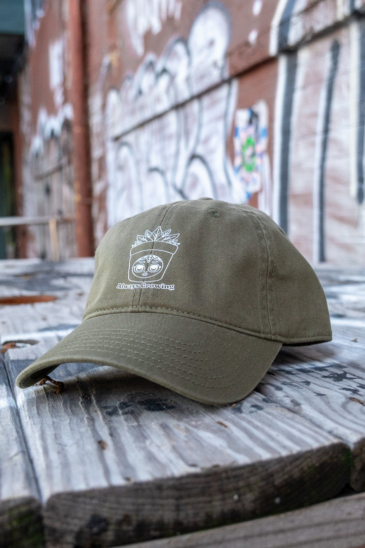 Olive green low-profile 'dad' cap with a white 'Always Growing' character logo, applied with DTF vinyl transfer, set against a wooden surface.