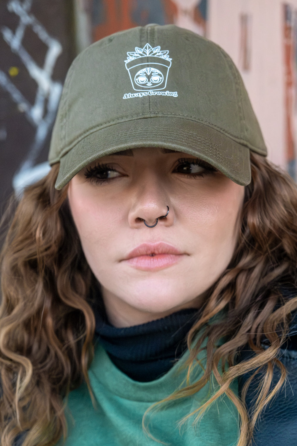 A woman with curly hair wearing an olive green 'dad' cap with a white 'Always Growing' DTF vinyl transfer logo, set against a soft-focus urban background.