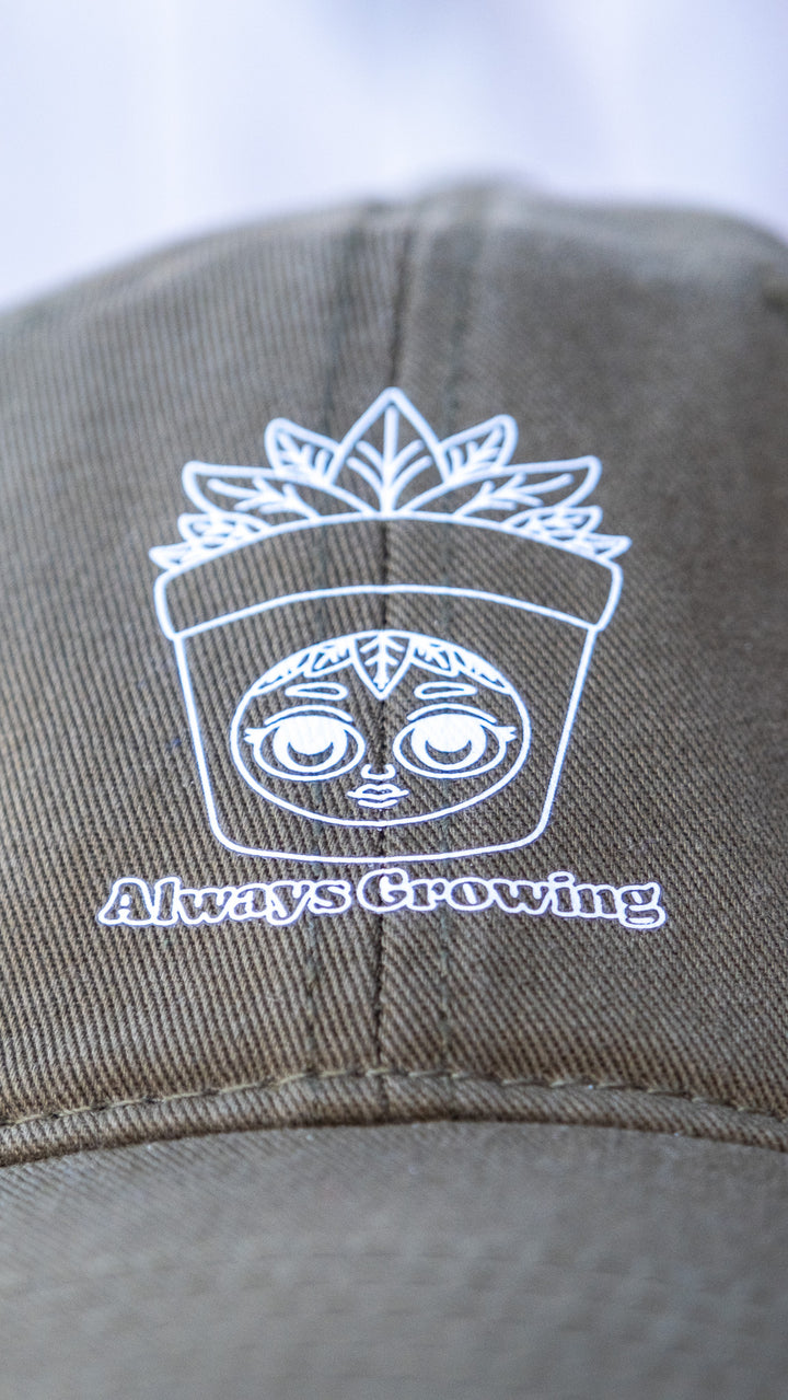 Close-up of the white 'Always Growing' logo on an olive green 'dad' cap, featuring a detailed DTF vinyl transfer of a whimsical face encased in a sprouting plant.