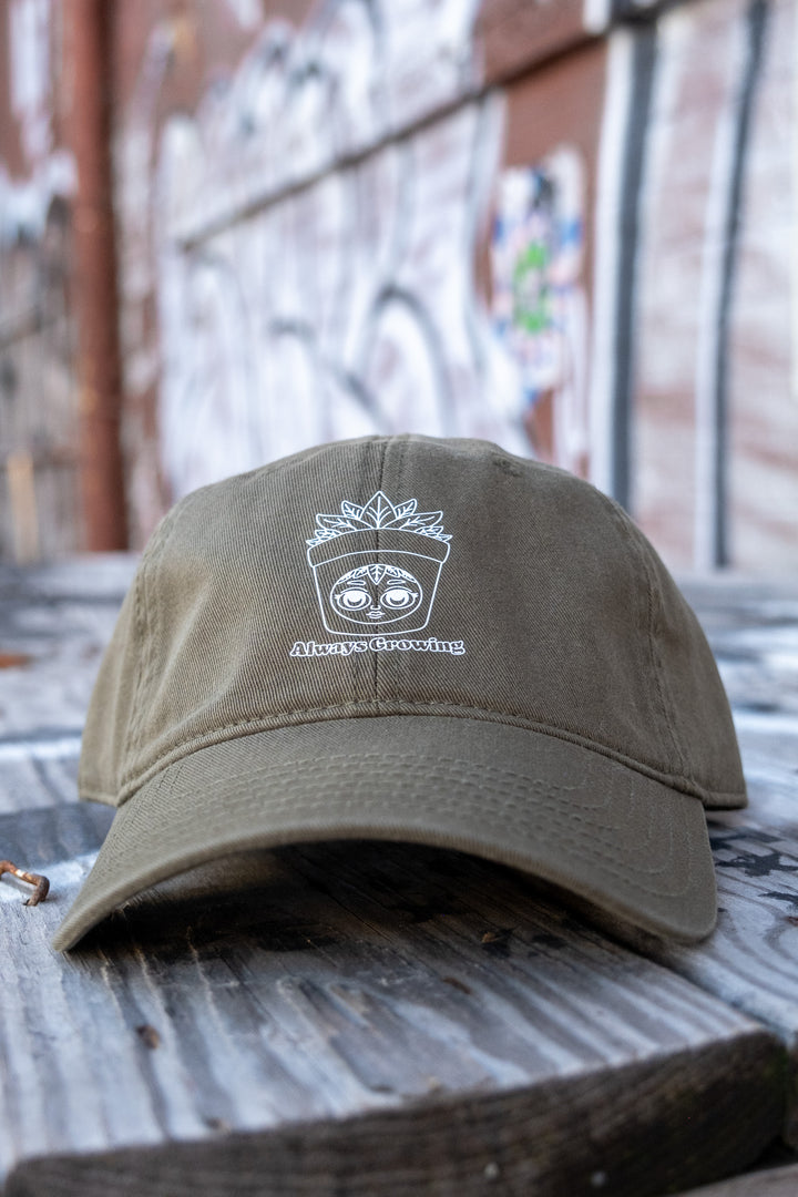 Olive green 'dad' cap with a detailed white 'Always Growing' logo, featuring a playful face and leaves design, showcased on a rustic wooden bench.