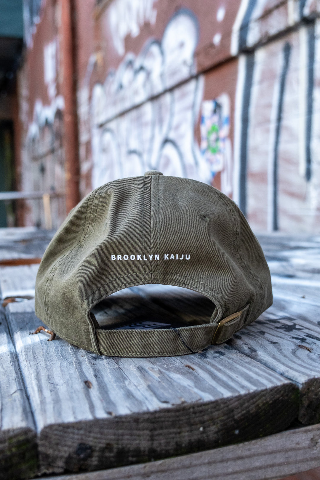 Rear view of an olive green 'dad' cap featuring 'BROOKLYN KAIJU' in bold above the adjustable strap, resting on an aged wooden bench with a graffiti backdrop.