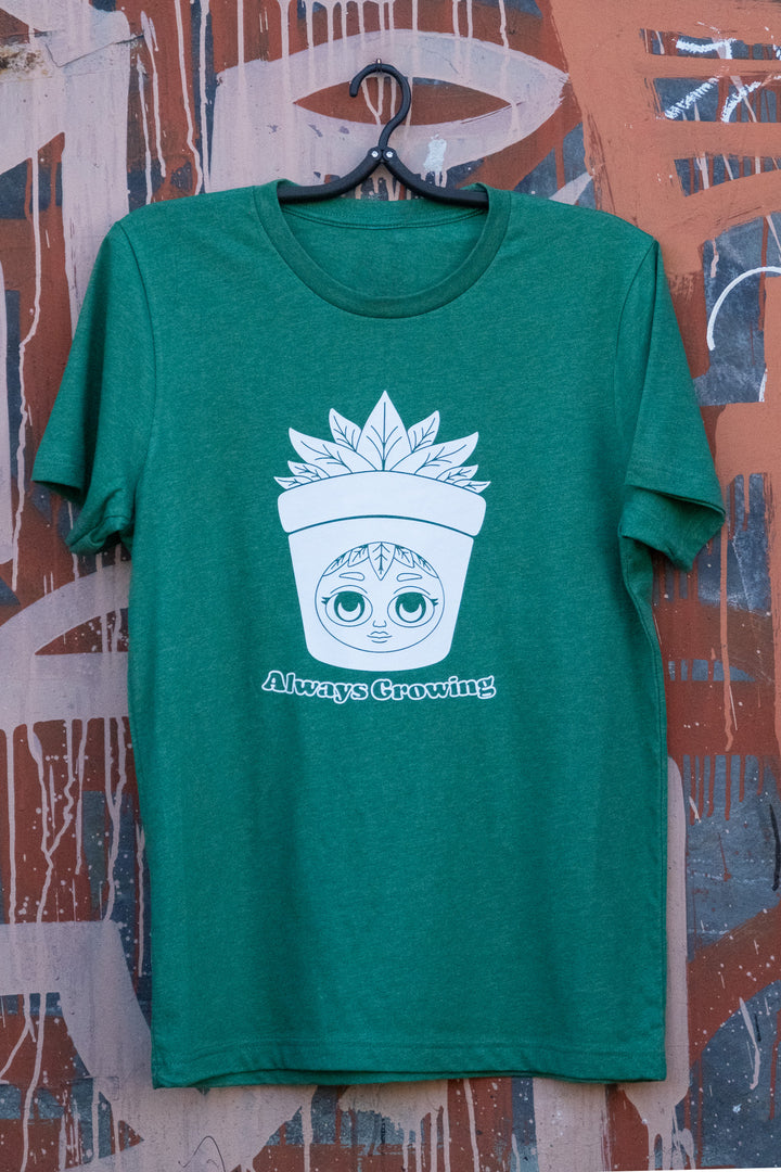 Forest green T-shirt with 'Always Growing' and a playful plant pot character graphic, displayed on a hanger against a weathered mural