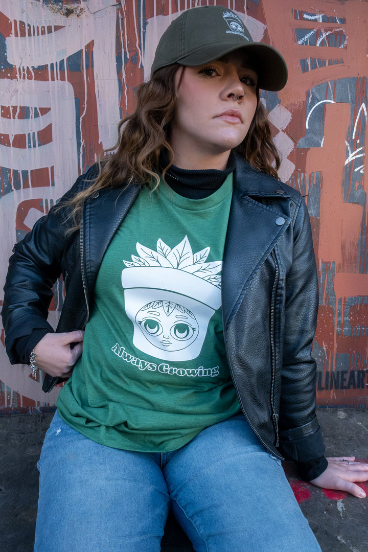 A casually styled woman in a green 'Always Growing' graphic tee, complemented by a muted green cap, stands against a graffitied wall.