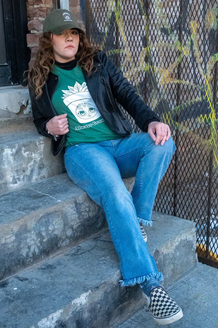 A laid-back vibe captured with a woman seated on steps in a green 'Always Growing' tee, denim jeans, and a cap, blending street style and comfort.