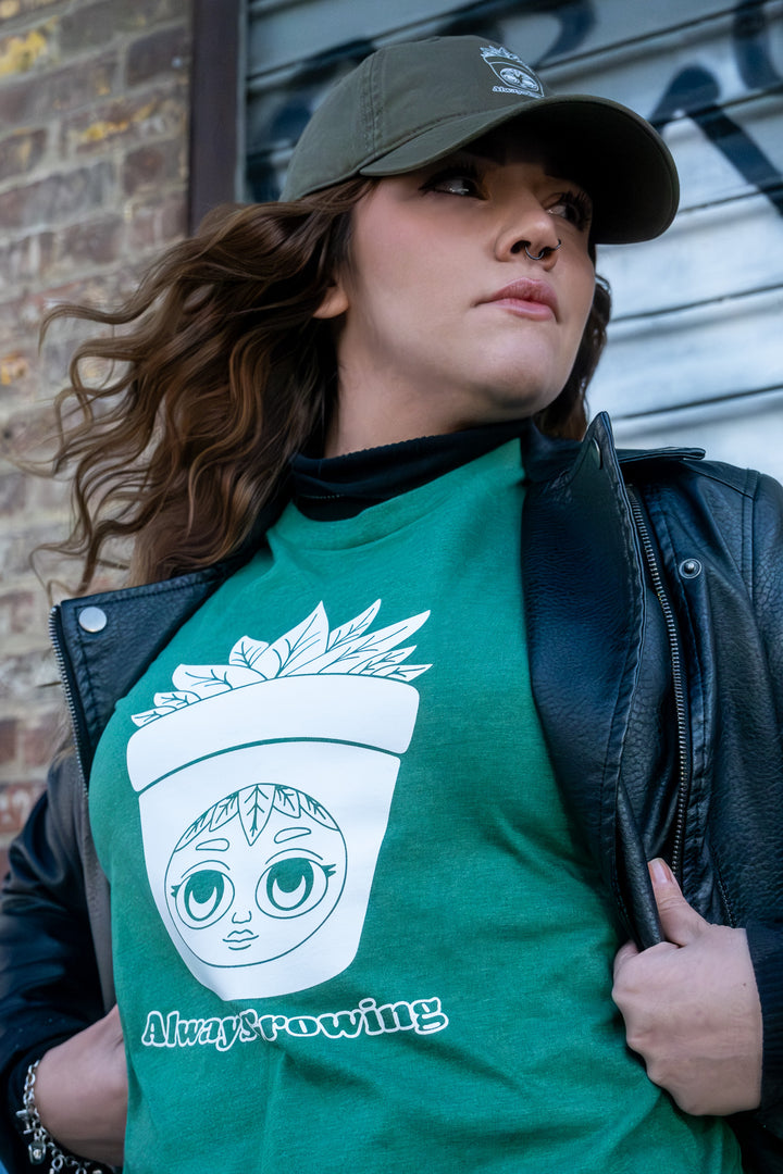 A close-up on a woman wearing an 'Always Growing' shirt, highlighting the whimsical pot plant character design, against a textured brick backdrop.