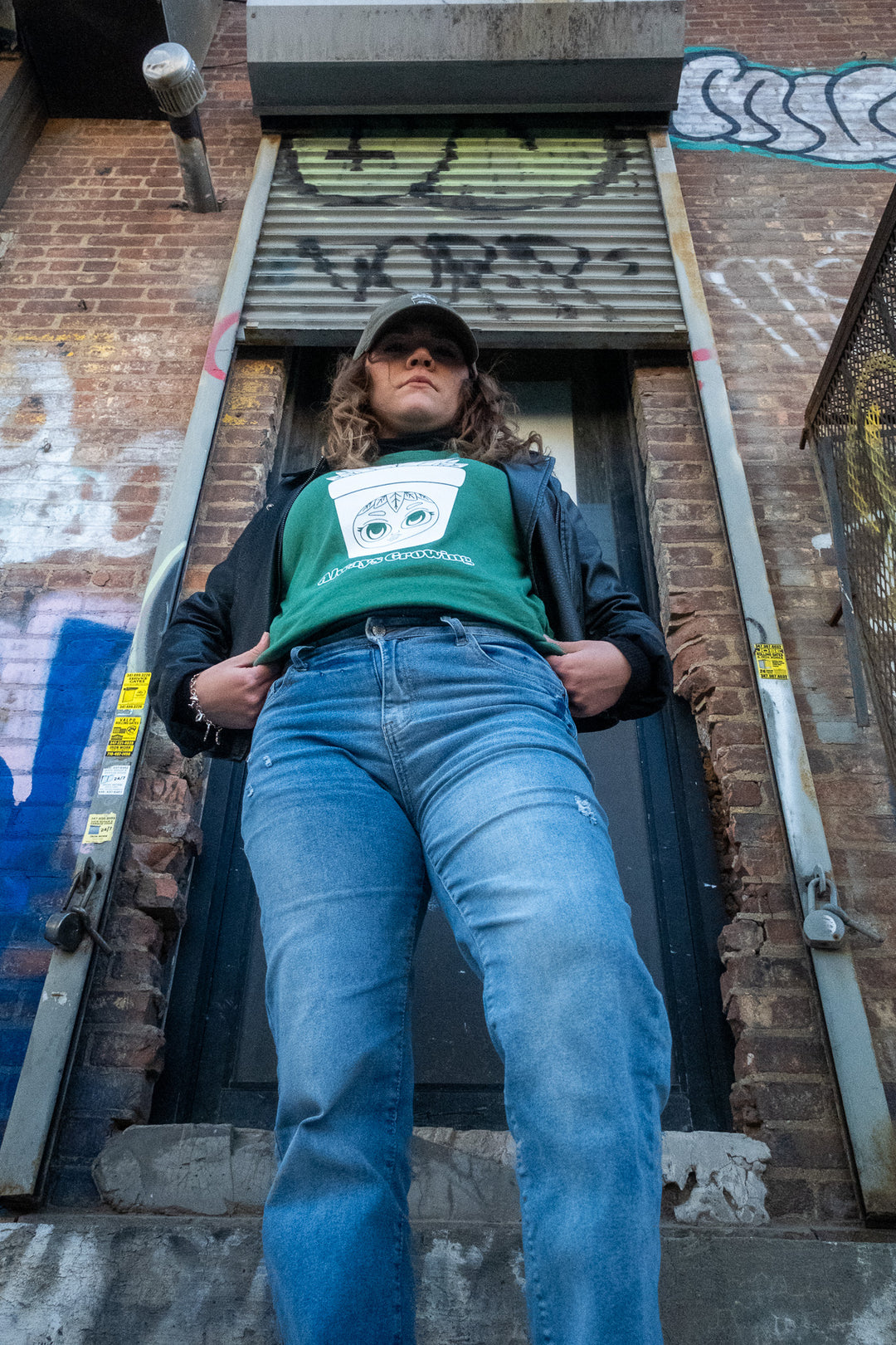 Woman in a dynamic pose wearing  a green 'Always Growing' T-shirt and cap, exuding confidence and street-smart style on a urban city building's stoop.