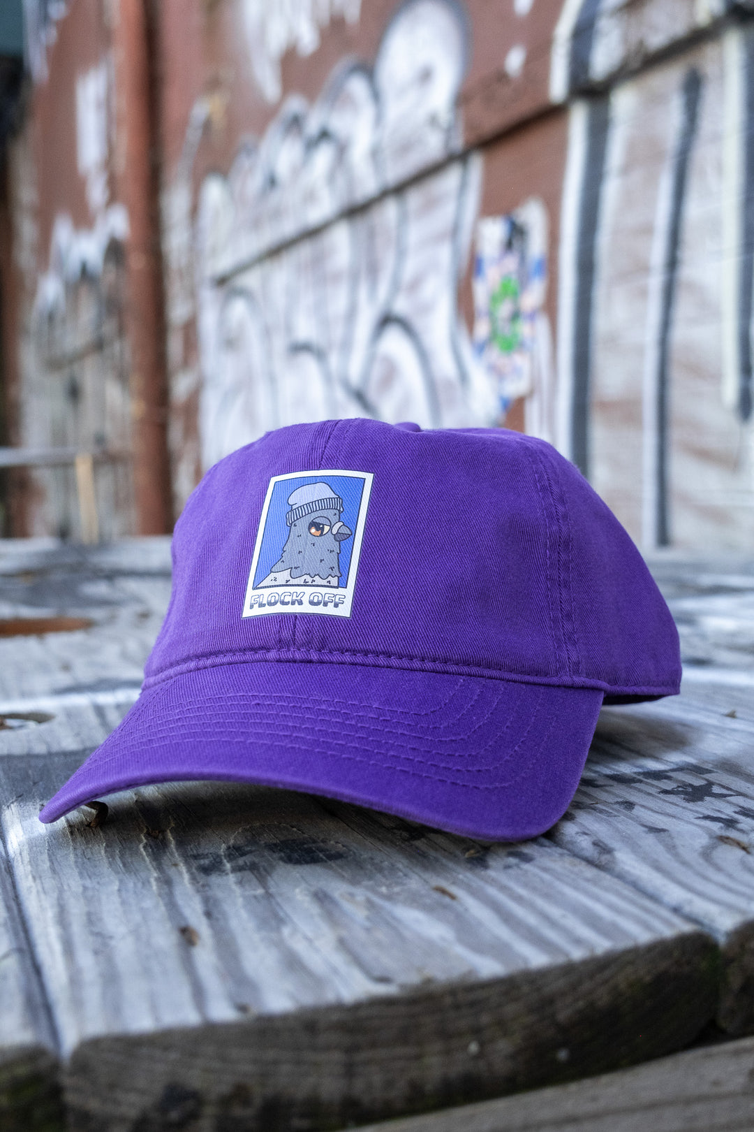 A purple low-profile 'dad' cap with a 'Flock Off' vinyl print centered on the front, placed on a weathered wooden bench.