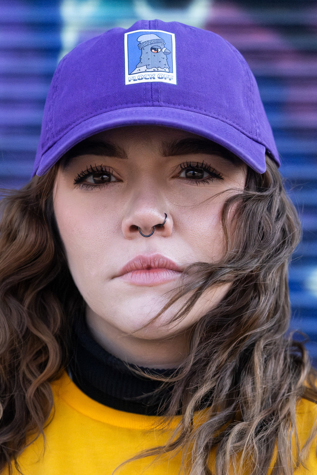 A woman wearing a purple 'dad' cap with a 'Flock Off' vinyl print, against a blurred multicolored striped background.