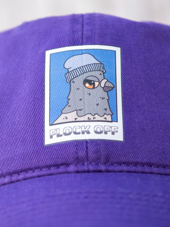 Close-up of a purple low-profile 'dad' cap's 'Flock Off' vinyl print, depicting a bird in a beanie with a stylized expression.