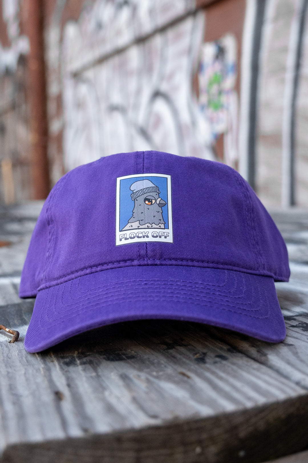 Purple low-profile 'dad' cap with a 'Flock Off' vinyl print displayed on a rustic bench, with a graffiti backdrop in soft focus