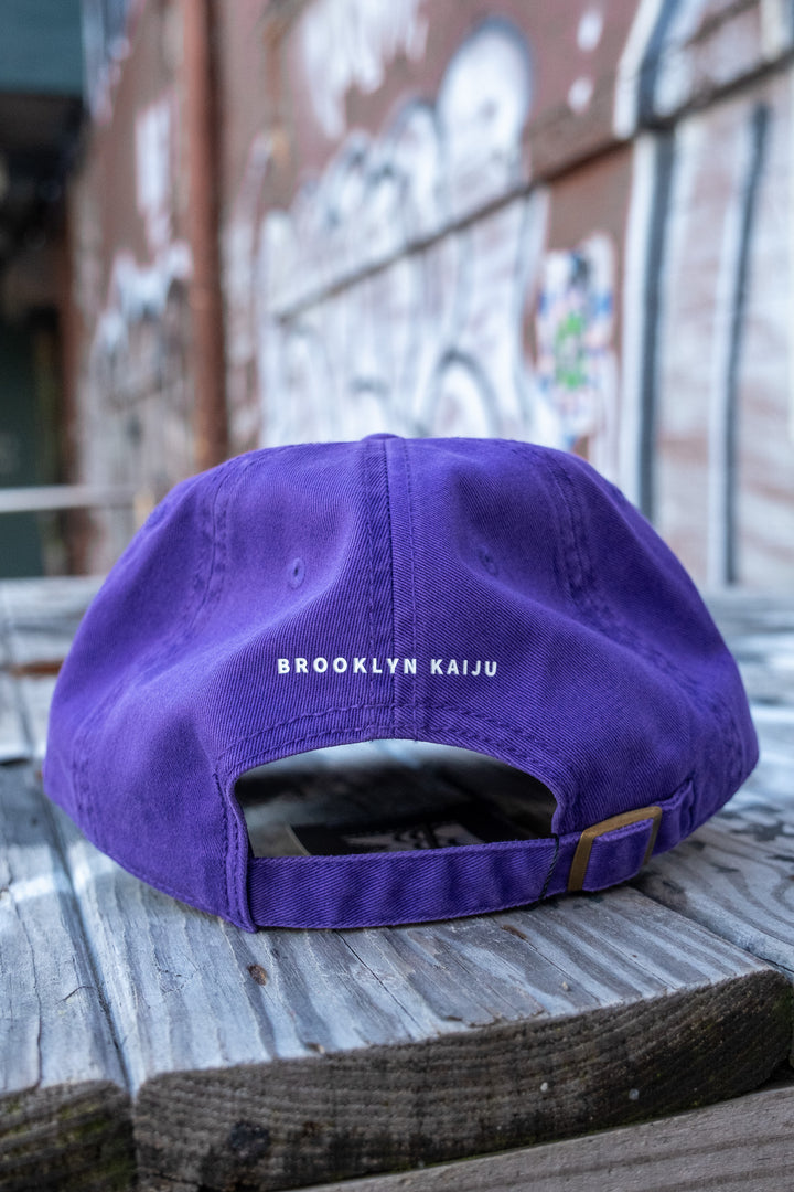 Rear view of a purple low-profile 'dad' cap showing the text 'BROOKLYN KAIJU' above the back strap, resting on a wooden bench.