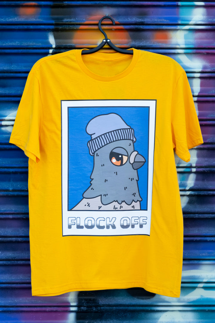 Vibrant yellow T-shirt featuring a quirky 'Flock Off' pigeon  in a knit cap design, hanging against an artistic spray-painted wall.