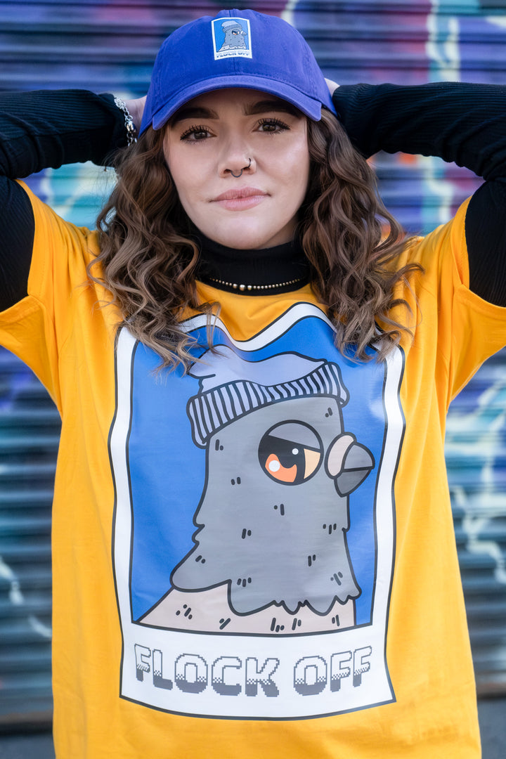 Confident woman in a yellow 'Flock Off' tee with a pigeon illustration, matched with a casual purple 'dad' cap, set against a dynamic graffiti wall