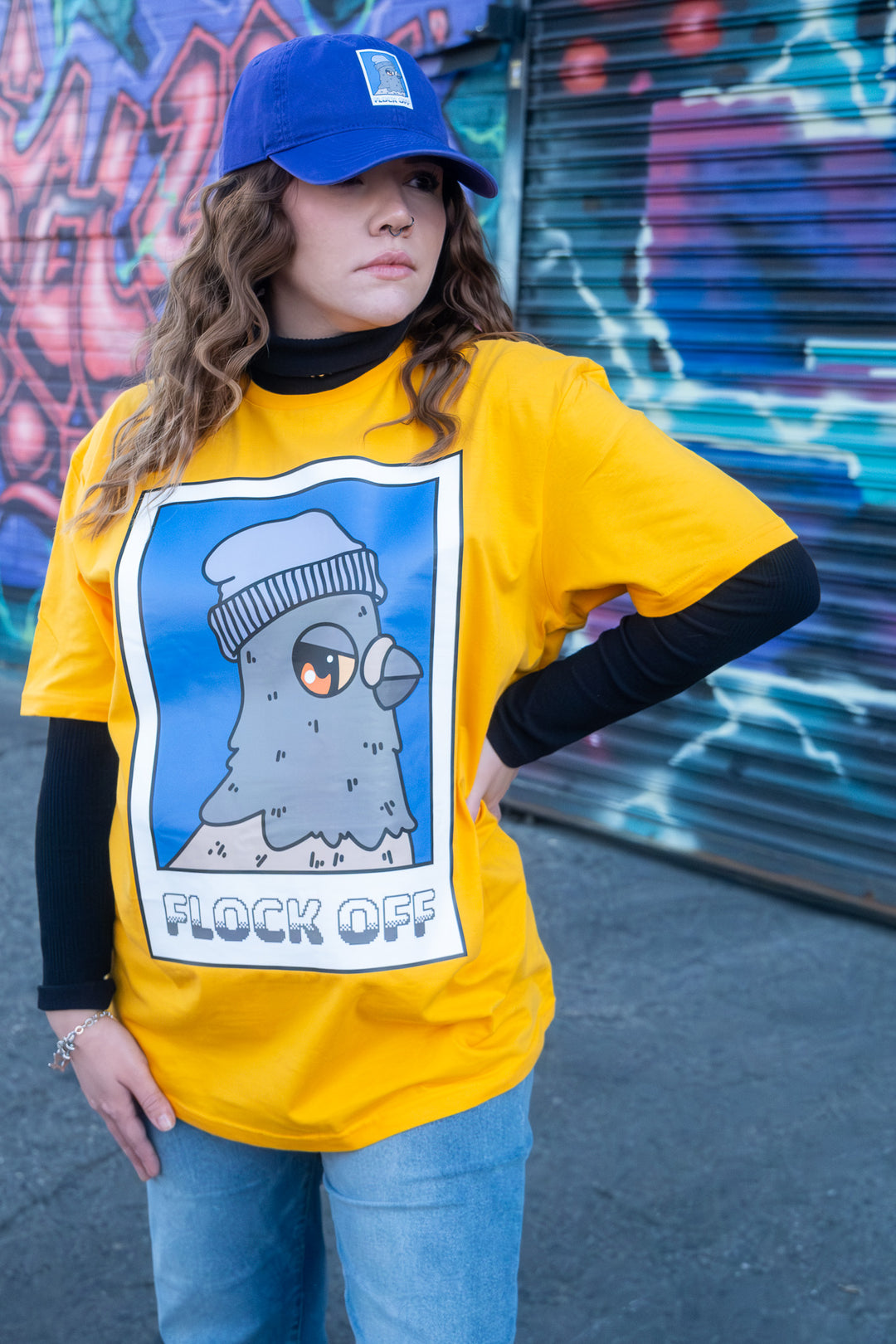 Model wearing a street style yellow T-shirt donning a 'Flock Off' beanie-wearing bird graphic, paired with denim jeans and a purple cap, amidst an urban backdrop.