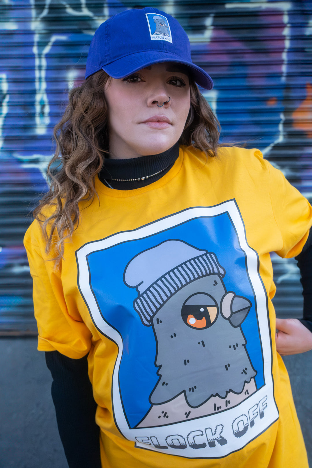 Stylish woman modeling a 'Flock Off' T-shirt with an eye-catching pigeon print, standing confidently with a blue cap, in an edgy graffiti-splashed alley.