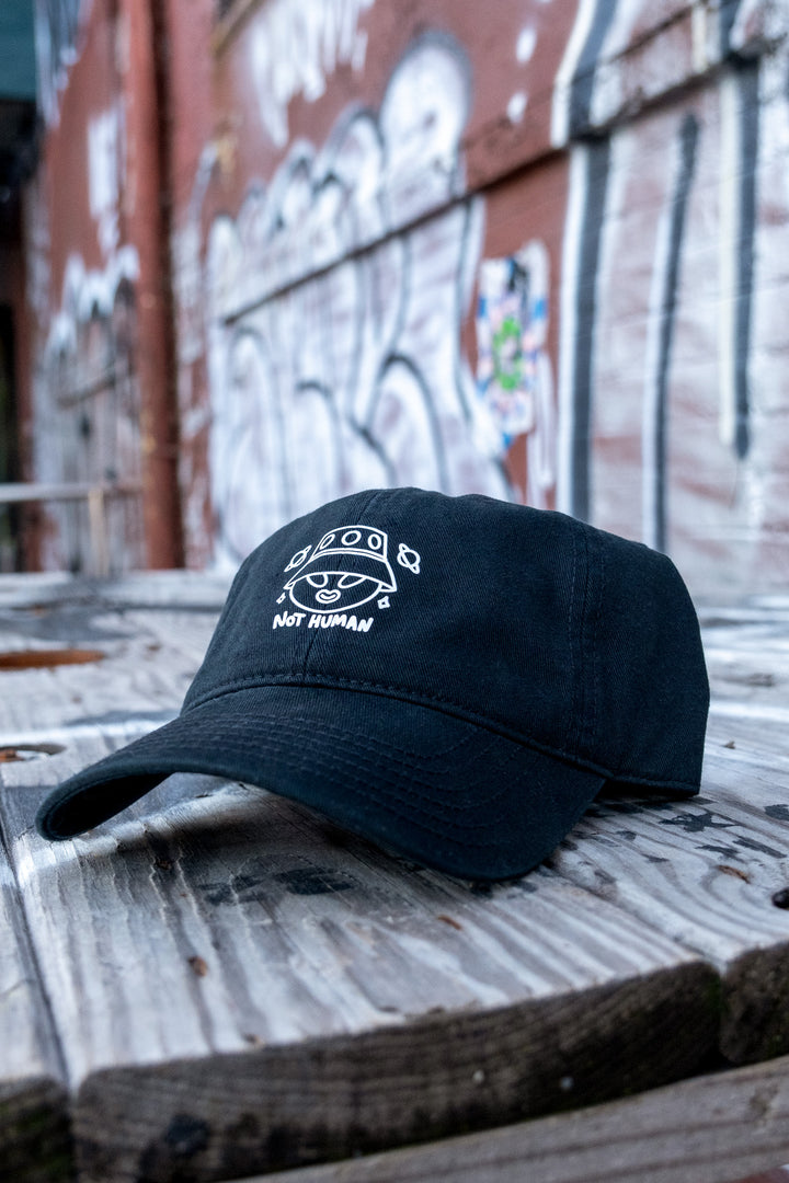 A black low-profile 'dad' cap featuring a white 'Not Human' vinyl print with an alien design, on a wooden bench.