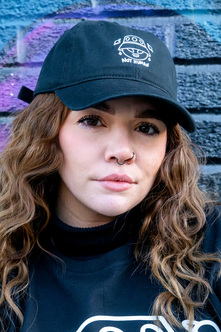 Black low-profile 'dad' cap with 'Not Human' white vinyl print, worn by a woman in front of a blurred graffiti wall.