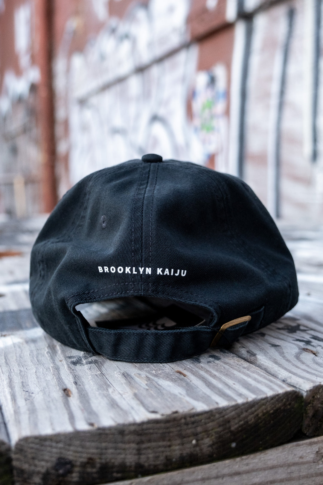 Rear view of a black low-profile 'dad' cap with 'BROOKLYN KAIJU' in white vinyl print above the strap, on a rustic bench.