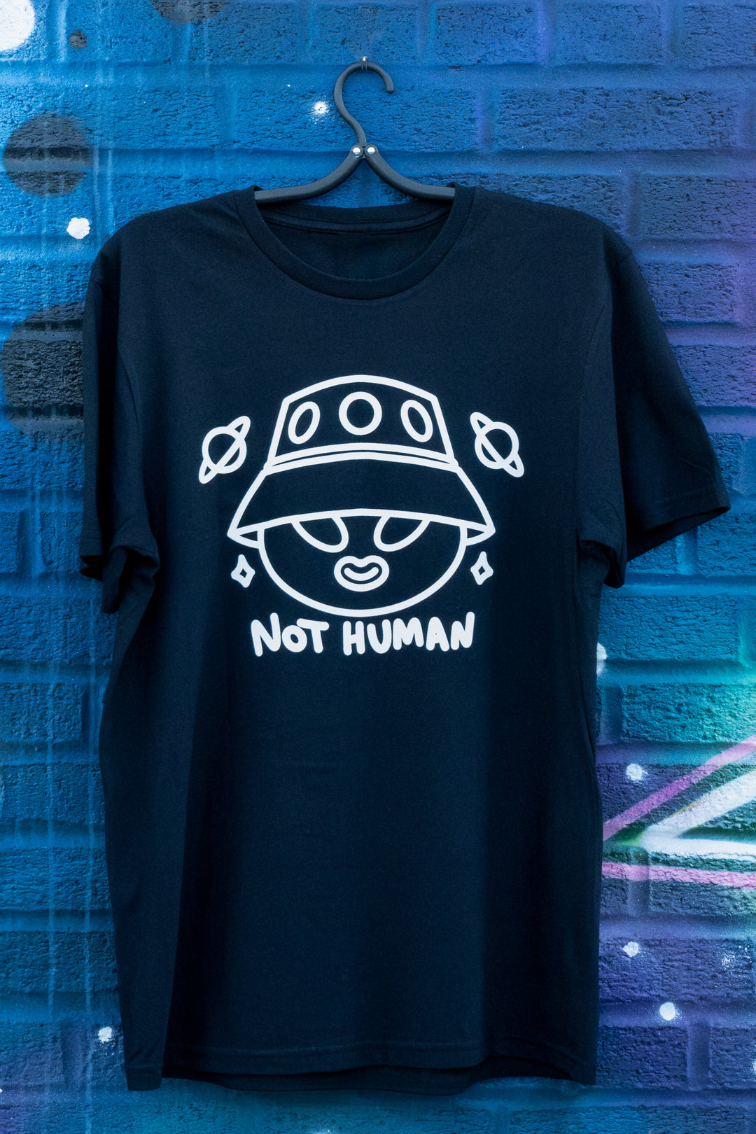 Black T-shirt with a quirky 'Not Human' alien design, displayed on a hanger against a creatively spray-painted blue brick wall.