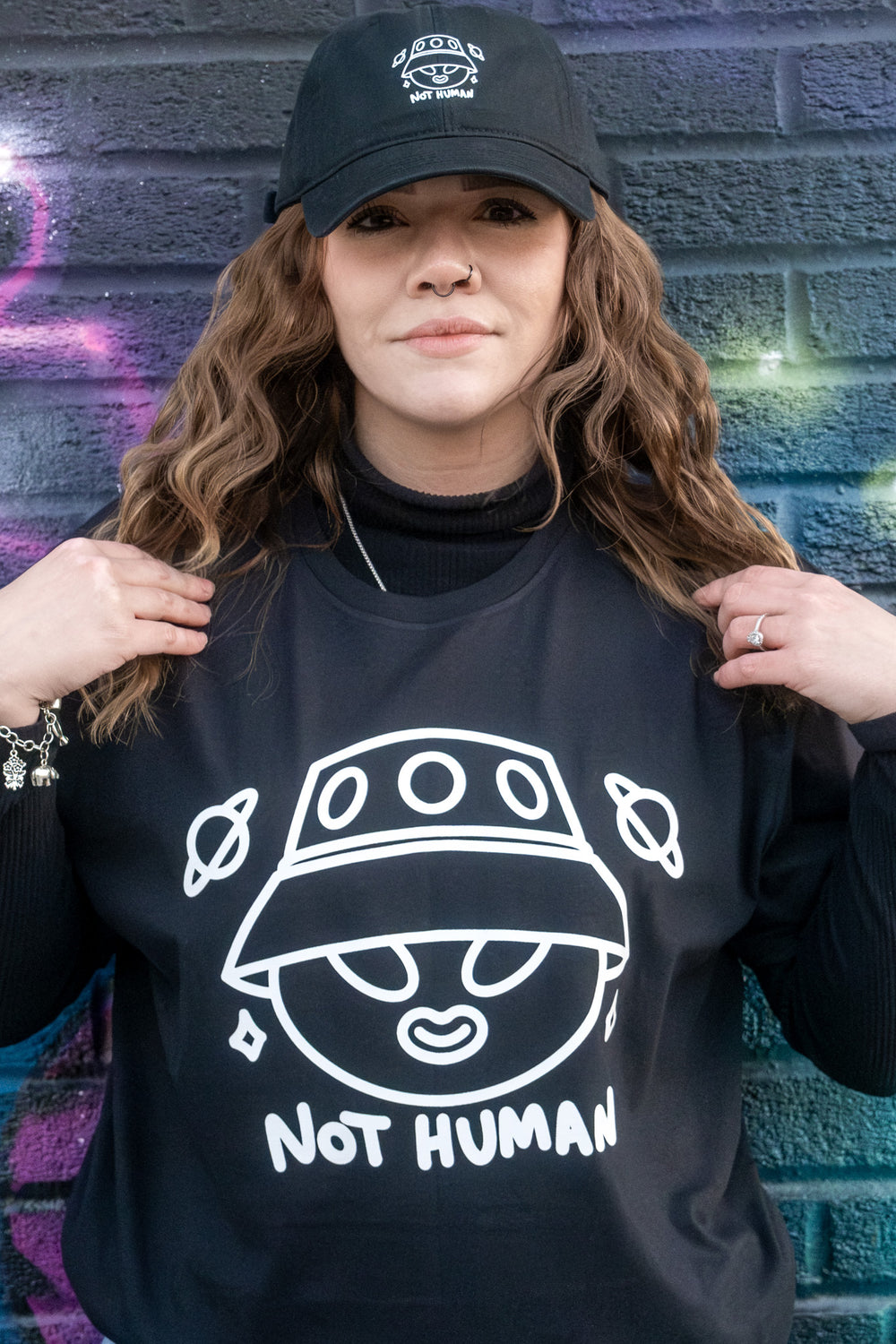 rendy woman showcasing a 'Not Human' alien print T-shirt paired with a black cap, complementing the colorful, graffiti art wall behind her.