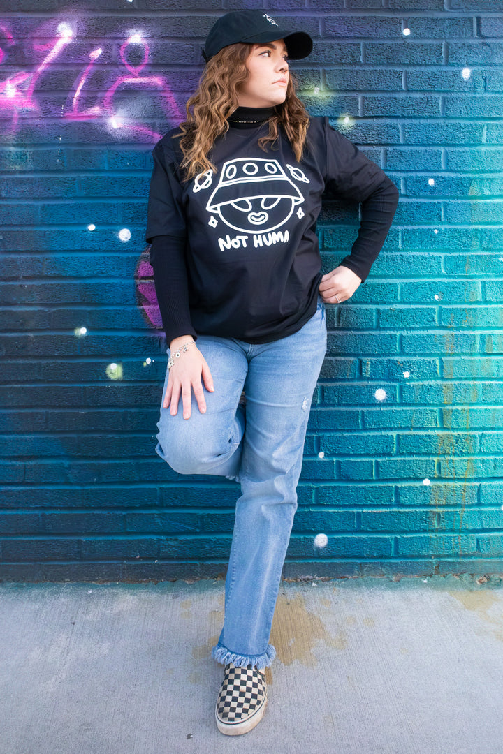 Street-style snapshot of a woman in a playful 'Not Human' T-shirt, denim jeans, and checkered sneakers, leaning on an urban wall.