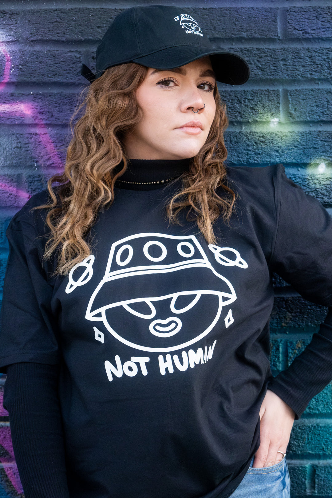 Confident stance of a woman modeling a 'Not Human' graphic tee, accentuated by a black cap, against a textured blue wall.