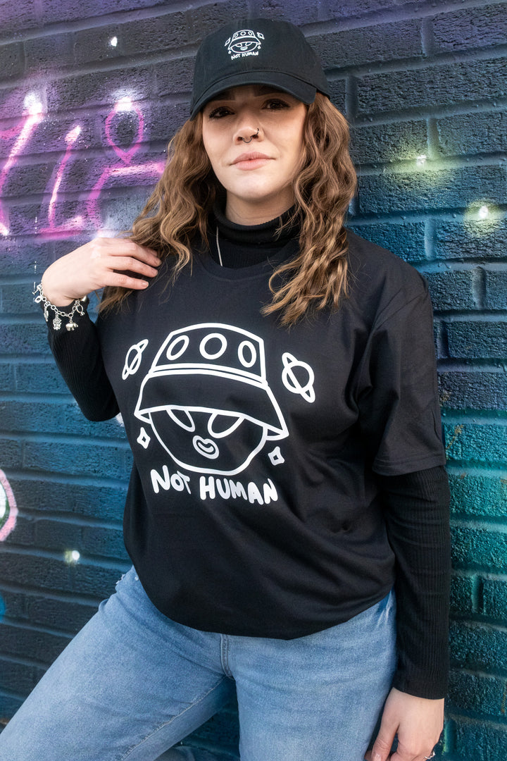 Casual and cool, a woman sports a 'Not Human' alien T-shirt and black cap, exuding relaxed streetwear vibes against an urban graffiti backdrop.